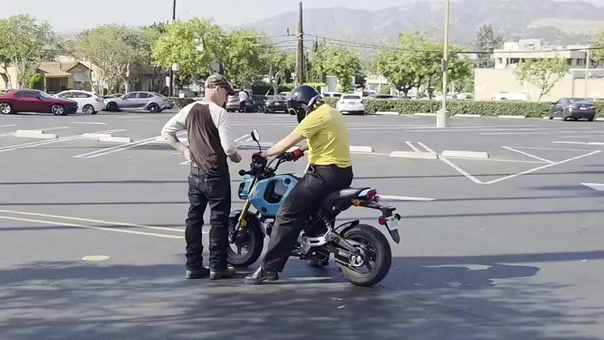 Two men are in a large, paved parking lot on a sunny day. The man on the left is wearing a baseball cap, a long-sleeved shirt, and dark pants. He is standing with his hands on his hips, looking at the man on the right. The man on the right is sitting on a small, blue motorcycle. He is wearing a helmet, a yellow t-shirt, and dark pants. The motorcycle is a street bike style. The parking lot has white lines marking parking spaces. In the background, there are trees, buildings, and cars parked. There are also mountains in the distance.