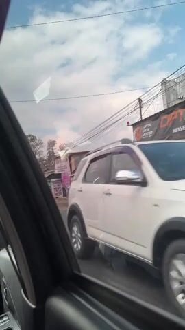 The video is filmed from inside a vehicle, looking out through the passenger-side window. A white SUV is visible in the foreground on the right, its rear tire in motion, indicating the vehicle is moving. Beyond the SUV, a street scene unfolds. A person wearing a pink shirt and pants walks away from the camera. In the background, there are several buildings, some appearing to be shops with signs. Power lines and utility poles crisscross the sky. The sky is partly cloudy with patches of blue. The overall environment appears to be an urban or semi-urban street.