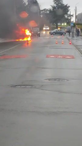 The video shows a car engulfed in flames in the middle of a street. Orange traffic cones are placed in a line to the right of the burning car. A group of people are standing on the sidewalk on the right side of the street, observing the fire. A white van with blue accents is parked on the left side of the street. The road is wet, and the sky is overcast. The fire is generating a lot of smoke. Red speed bumps are visible on the road.