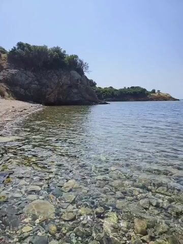 A video shows a view of a clear, shallow ocean. The seabed is covered in colorful pebbles. A rocky cliff covered in green foliage rises from the left side of the frame. In the distance, a landmass can be seen, also covered in green foliage. The sky is a clear blue.