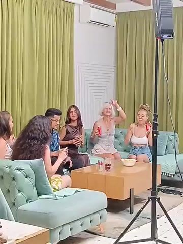 The video shows a group of six people sitting on a teal-colored sofa and chairs in what appears to be a living room. They are holding cans and glasses, seemingly enjoying themselves. A man approaches the group with a camera and takes pictures of them. In front of the group is a light-colored wooden coffee table with drinks and a bowl of snacks. The room is decorated with green curtains and a geometric design on the wall. A light stand is positioned on the right side of the frame, providing additional lighting.