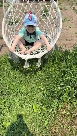 A young boy, wearing a light blue t-shirt, black shorts, and a blue baseball cap with a Spider-Man design, sits in a white macrame hanging chair outdoors. The boy is looking down and smiling. The chair is suspended by ropes from what appears to be a wooden structure. The setting is grassy with some dry dirt patches and scattered green plants. Sunlight suggests it is daytime, likely a warm day given the outdoor setting and the boy's attire. The scene appears to be a backyard or a similar outdoor recreational area. The boy is holding onto the sides of the chair. The bottom edge of a person's hand with white fingernails is visible in the foreground, suggesting the recording person is interacting with the boy. The cap has the word "SUPER" visible on it.