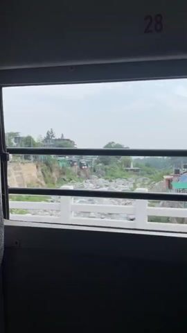 The video is a first-person perspective shot from inside a vehicle, looking out a window. The window is framed by dark grey interior panels. Outside the window, there are several horizontal black bars that appear to be part of the vehicle's structure or possibly window bars. Beyond these bars, a white railing is visible, with a river flowing beneath it. The riverbed is rocky, and there are trees and buildings on the far bank of the river, suggesting a rural or semi-urban setting. The sky is overcast with some clouds. The numbers "28" and "29" are visible in red at the top of the window frame, likely indicating seat numbers. The camera appears to be in motion, as evidenced by the slight blur and the changing perspective of the landscape.
