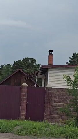 The video pans across a residential area with a cloudy sky. There are fences made of corrugated metal and wood, a brick pillar, a large metal barrel with a white bag next to it, a black minivan, and a wooden building with a slanted roof. Trees, including pine trees, are visible in the background, along with houses with red and green siding and red roofs. There are also utility poles with some signage. The overall scene depicts a rural or suburban setting.
