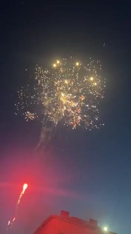 The video shows fireworks in the sky during the night. The fireworks are red and yellow. The sky is dark blue, with some light at the bottom. Some buildings are visible at the bottom of the video, and a street light is on the bottom right.