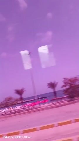 This video appears to be captured from a vehicle moving along a coastal road in Caraballeda, Venezuela. The scene is dominated by a vibrant purple filter, giving the sky, water, and even the sand a surreal, dreamlike quality. 

The primary subjects are several palm trees lining a beach. Beyond the trees, the ocean stretches out, meeting a rocky shoreline. The beach itself is dotted with numerous pink umbrellas, suggesting a public beach area. In the foreground, a road with yellow markings is visible, reinforcing the idea that the footage is taken from a moving vehicle. A few buildings, possibly small shops or beachside facilities, are also present. A dark-colored SUV is parked near the roadside in one frame.

The activity taking place is a scenic drive along the coast. There are no people clearly visible in the footage, but the presence of beach umbrellas implies human activity. The time of day seems to be daytime, given the bright sky, though the purple filter obscures natural lighting cues. The weather appears to be clear, with some clouds visible.

Notable details include the pervasive purple color cast, the presence of palm trees and beach umbrellas typical of a tropical coastal setting, and the road markings indicating a driving perspective. The overall mood evoked by the filtering is one of tranquility and a slightly surreal, dreamlike atmosphere.

The visible text at the bottom of the screen reads "Infinix HOT 50Pro," which is likely the model of the phone used to record the video.