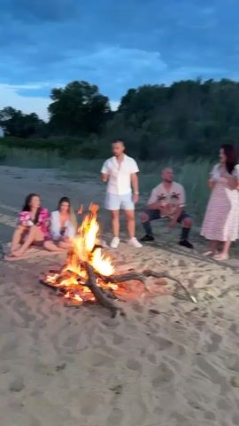 The video shows a group of people gathered around a campfire on a beach. There are four women and two men. One man is standing while holding a glass, another is sitting. Two women are seated in chairs. The other two are seated on a blanket on the sand. The campfire is burning brightly in the center of the group. The beach is sandy, and there are trees and grass in the background. The sky is cloudy. People appear to be drinking and talking. The camera moves closer to the fire and then back out to the group.