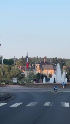 The video captures a view of a roundabout with a fountain in the center. A French flag flies on a flagpole to the left of the fountain. In the background, there are buildings with pitched roofs, trees, and a clear sky. Traffic signals and road markings, including a pedestrian crossing, are visible in the foreground. The fountain is active, with water spraying upwards. The sun appears to be low, casting a warm light on the scene. The camera is static for most of the clip, but there is a slight pan to the right towards the end.