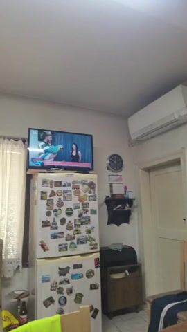 A television is mounted on top of a refrigerator in a room. The television displays a program with two people. The refrigerator is covered in numerous stickers. To the right of the refrigerator, a clock is mounted on the wall. Below the clock, a small shelf holds various items. To the right of the shelf, a door is visible. An air conditioning unit is mounted on the wall above the door. A window with curtains is visible to the left of the television. The room appears to be a kitchen or dining area.