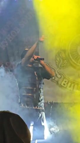 A rapper performs in front of a black and white banner. The banner features a motorcycle wheel, wings, and the words "The Riders Bulgaria MC." The stage is lit with blue, yellow, and green spotlights. The rapper wears a dark outfit and a baseball cap, and performs with a microphone. There is a large amount of fog or smoke on stage.