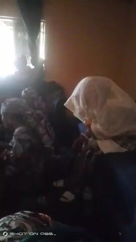 The video shows a dimly lit interior room with about seven people, mostly women, seated or positioned low to the ground. Light streams in from a window, creating overexposure in that area. The figures appear to be wearing patterned clothing.  Some have head coverings. The exact nature of their activity or interaction isn’t clear due to the low lighting and lack of clarity. The phrase "SHOT ON P65_" is displayed at the bottom left of the frame.