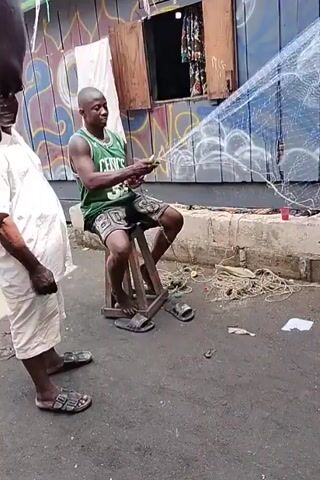 Here is a concise description of the video:

The video shows a man with a striped shirt walking toward another man sitting on a ledge near a building, potentially a storefront, that has some graffiti. The man is repairing or working on a large fishing net. There is some rope and other debris on the ground. The video is shaky, as though it was taken while the camera was moving.