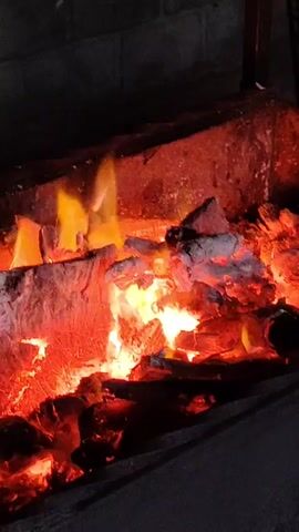 The video shows a close-up shot of a burning fireplace. Logs and embers glow with intense red and orange light. Flames flicker and dance above the wood. The background is dark, suggesting it is nighttime or the surrounding area is unlit. A portion of a dark, possibly metal or stone, structure frames the fireplace. There is no visible activity or interaction, only the natural process of burning wood.