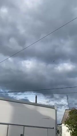 The video shows a military jet flying rapidly across a heavily overcast sky. A loud, sharp sound, characteristic of a jet engine, is heard immediately at the start of the recording, indicating the jet is passing by at high speed. The aircraft appears as a small, dark silhouette against the grey and white clouds, leaving a faint exhaust trail behind it as it moves quickly from left to right and away from the camera. In the foreground, power lines stretch across the upper part of the frame, and the edge of a white building or structure along with a wooden utility pole are visible below, suggesting an urban or suburban setting. The loud jet sound quickly diminishes as the aircraft disappears into the distance. The location context provided is Telma, Russia.