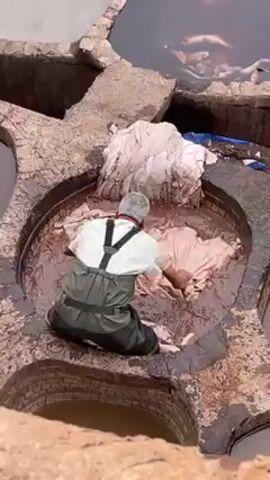A middle aged man kneels waist-deep in a circular vat containing a brown liquid. The man wears waterproof waders and a light-colored short-sleeved shirt. He pushes a large pile of light-colored leather or fabric into the vat and manipulates it with his hands. The vat is surrounded by other similar vats. The vats are constructed with bricks and concrete, appearing aged and weathered.  The area outside the vats looks to be a work area. The view is from an elevated position looking down.