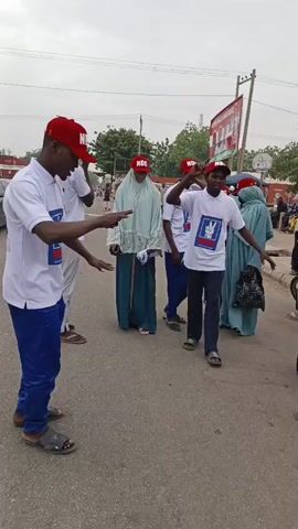 The video captures a lively political gathering on a city street. Several individuals wearing white polo shirts and red baseball caps with "NDC" emblazoned on them are dancing and gesturing animatedly. The shirts also feature a political logo and a portrait on the back. Some participants are dressed in traditional Muslim attire. The street is paved and lined with buildings, trees, and power lines. There are vehicles, including a tricycle, visible in the background, suggesting a public space with regular traffic. The atmosphere appears celebratory and energetic, indicative of a political rally or campaign event.