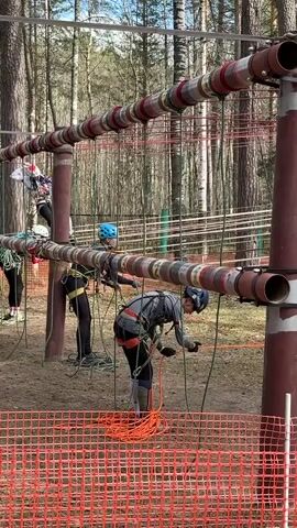 The video shows several people participating in an outdoor rope course in a wooded area. The course is constructed of brown metal poles and pipes. The people are wearing safety harnesses and helmets, and they appear to be securing or adjusting their ropes. An orange safety fence is visible in the foreground, indicating the perimeter of the course. The area has a sandy ground with tall trees surrounding the course. The sky is visible through the trees, suggesting a sunny day.