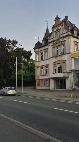 A video captures an old, ornate building with a grey facade, white trim, and several windows. The building has a distinct turret on the left and a decorative facade on the right. Adjacent to the building is a modern structure with a yellow awning and large glass windows, possibly a storefront. In the foreground, a grey asphalt road with white lane markings is visible. Trees line the left side of the road, and a single streetlamp stands tall. A crescent moon is visible in the twilight sky. A dark-colored car is seen driving away from the camera on the road. The overall scene suggests an urban or suburban setting during dusk.