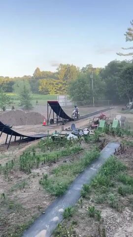 A person on a dirt bike jumps off a ramp and into the air. The dirt bike is white and black, and the rider is wearing a helmet and protective gear. The rider is in mid-air, and the dirt bike is angled upwards. The background is a blue sky and trees. The video then shows the dirt bike landing on an inflatable ramp. The rider is safely on the ramp. The environment is a dirt track with ramps and jumps. There are trees and grass in the background.