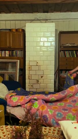 A man is seen in a static shot of an indoor setting. He is quickly throwing a patterned blanket over a partially visible person who is reclined on a blue couch. The couch is laden with cushions and a floral-patterned blanket. The person under the blanket has their legs stretched out. The room contains a brick structure and bookshelves filled with books. The scene is lit by an unseen light source.