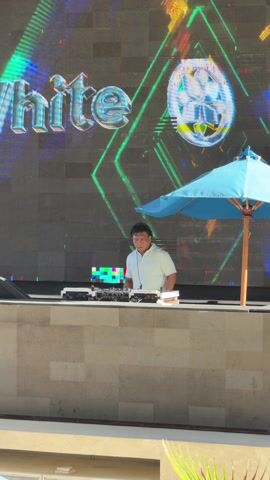 A DJ is performing at an outdoor venue. He is standing behind a low wall, facing the camera, with his hands on the DJ equipment. He is wearing a light-colored button-down shirt. Behind him is a large LED screen displaying animated graphics and the word "White". To the right of the DJ is a blue beach umbrella. The scene is well-lit, suggesting it is daytime.