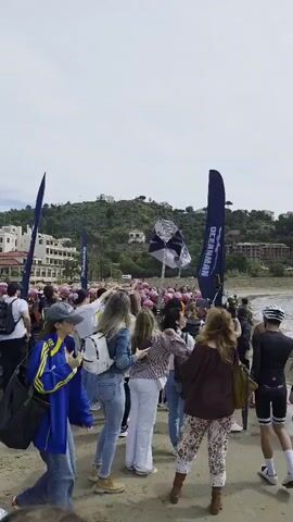 A crowd of people gathers on a sandy beach under a partly cloudy sky. Many wear pink swim caps, suggesting an organized event related to swimming or water sports. Blue flags with a logo are positioned throughout the crowd and near a large black archway, likely marking the start or finish line. The crowd seems to be gathering or waiting for an event to begin, with many holding phones and taking photos. In the background, there are buildings along a hillside, adding to the scenic coastal setting. The general atmosphere is lively and expectant.