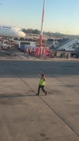 A person wearing a high-visibility vest and headphones walks across an airport tarmac. They are holding two red batons, one in each hand, which they are gesturing with. In the background, there is an airplane, a set of stairs with red and white striped markings, and various other airport equipment. The sun is casting long shadows on the ground.