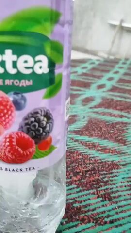A close-up shot focuses on a bottle of iced tea with a label depicting various berries, including raspberries, blueberries, and blackberries. The bottle appears to be filled with a clear liquid and condensation. The background is blurry, but a textured surface, possibly a table or cloth, with a pattern of red and green can be seen. The lighting suggests an indoor environment, possibly natural light.