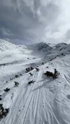 The video shows a snowy mountain landscape with a few buildings in the distance. The sky is cloudy. The foreground is covered in snow with some small patches of vegetation. Ski tracks are visible in the snow. The camera pans slightly to the left, providing a wider view of the mountainous area and the buildings nestled within the snow-covered valley.
