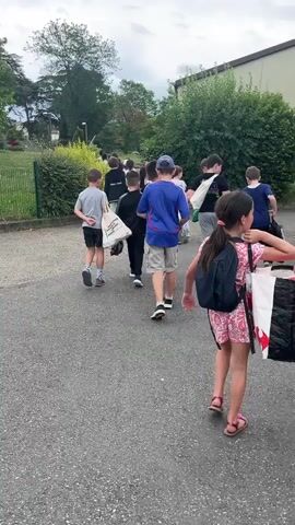 A group of children are walking away from the camera down a paved path. The children are of varying ages and appear to be walking in single file or in small groups. Some of the children are carrying backpacks or bags. The path is lined with bushes and trees on the left side, and a building is visible on the right side. The sky is overcast.