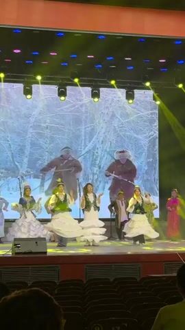 A group of performers is on a stage, engaging in a vibrant dance performance. Several women in long, flowing white dresses with green vests are dancing in the center, performing synchronized movements with raised arms. Other performers, including men and women in various traditional-looking costumes, are also on stage, some moving and others standing.

In the background, a large LED screen displays dynamic scenery. It transitions from a winter forest scene with two large figures, to a snowy landscape with wild horses running, and then to a close-up of a person riding a horse. The stage is brightly lit with colorful spotlights, primarily yellow, purple, and green, enhancing the theatrical atmosphere.

The video is filmed from an audience perspective, showing the backs of audience members' heads in the foreground, seated in rows of theater seats. The overall scene suggests a cultural or folk performance, possibly with storytelling elements, given the changing background visuals.