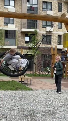 A girl is on a circular rope swing with a mesh seat. She is wearing a grey t-shirt and grey pants. She is swinging back and forth. A woman in a dark green jacket and dark pants is standing to the right of the swing, watching the girl. The playground has a gravel surface. In the background, there is a modern apartment building. Another child is visible on a climbing structure in the background.