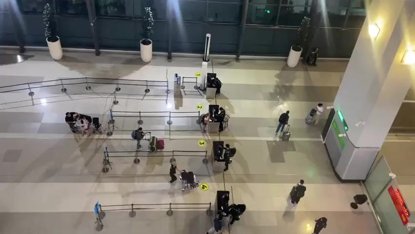 The video provides an overhead view of an airport terminal. Several people are visible, most of whom are pulling luggage. They are queuing at what appear to be check-in or security counters. Barriers are in place to organize the queues. Some individuals are standing, while others are seated in wheelchairs. There are also staff members at the counters, interacting with the travelers. The environment is indoors with tiled flooring and large windows in the background. Lighting fixtures are visible on the ceiling and walls. Two large potted plants are also present in the scene. The overall atmosphere appears to be one of routine travel operations.