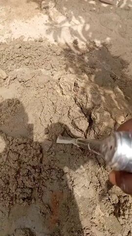 A shovel is used to mix what appears to be cement on the ground. There is a bag of cement in the background. The ground is dirt-covered and uneven. There is also some shade in the video, likely from a tree. The person operating the shovel is not visible.