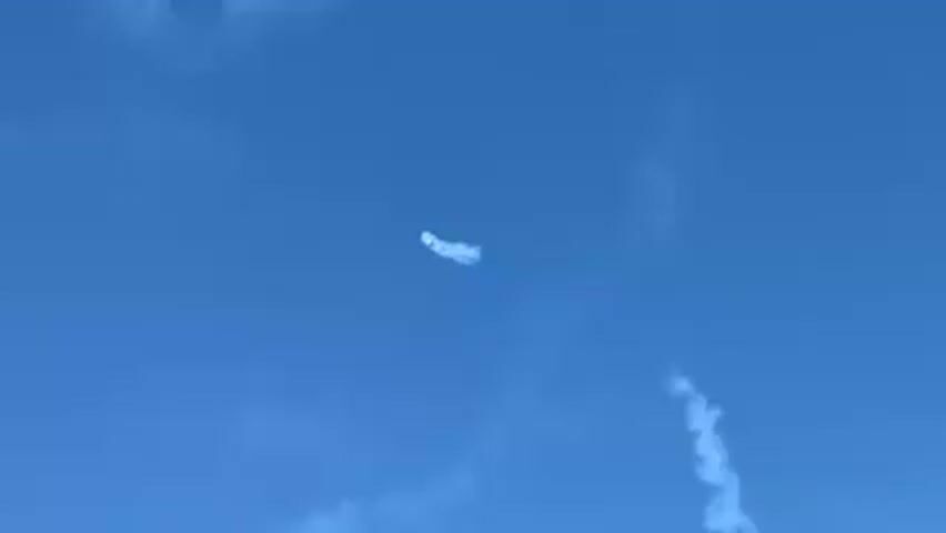 A small airplane performs an aerobatic maneuver in a clear blue sky, emitting white smoke. The smoke trails behind the plane as it rotates and descends. Other faint contrails are visible in the background.