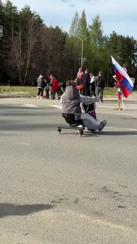 Two children ride wheeled office chairs down an asphalt road, propelling themselves with their feet, passing by a group of people standing on the side of the road by a Russian flag. There is a line of trees in the background.