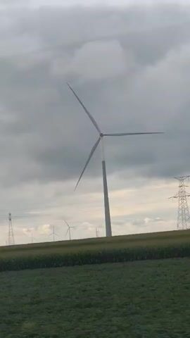 The video shows a landscape with wind turbines and agricultural fields under a cloudy sky. The camera appears to be moving, possibly from a vehicle, as indicated by the motion blur in the foreground. Several tall wind turbines are visible, with one prominent turbine in the center-left of the frame. In the background, there are power lines supported by a tall transmission tower. The fields consist of green crops, likely corn, with rows of plants visible. The overall scene depicts a rural area with renewable energy infrastructure and agriculture.