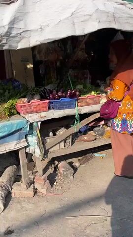 A woman wearing a brown hijab and an orange shirt with a floral pattern is standing in front of a stall. She is facing away from the camera. The stall appears to be selling various items, including packaged goods and what look like dried foods. There are also some terracotta-colored plates stacked to the right of the stall. A blue plastic bin is visible on the ground in front of the stall. Part of a red motorcycle is in the foreground. The ground is dirt or gravel, and the lighting suggests it is daytime with sunlight.