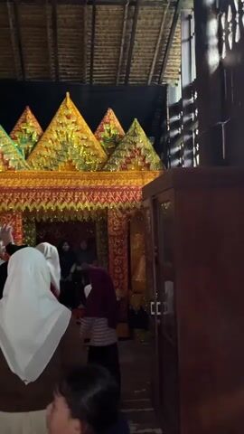 The video shows a group of people in what appears to be a traditional wooden building. In the background, there is an ornate, gold and red structure that resembles a stage or altar, decorated with triangular shapes. Several people are walking towards this structure, some of whom are wearing hijabs. To the right, there is a dark wooden cabinet with glass doors displaying items inside. The walls of the building are made of wood, and there is a framed photograph on one of the walls. A window with wooden shutters is open, revealing a dark interior. The people in the foreground are facing away from the camera, and their clothing suggests a casual setting. The lighting is dim, with some areas brightly lit by the decorations in the background.