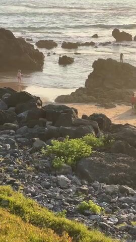 The video shows a rocky coastline with several large boulders. The ocean is visible in the background with gentle waves. There are several people on the beach and in the shallow water. One person is standing in the water, another is walking on the sand, and two people are on a large rock formation. The sun appears to be setting, casting a warm glow on the scene. The foreground is covered with grass and smaller rocks.