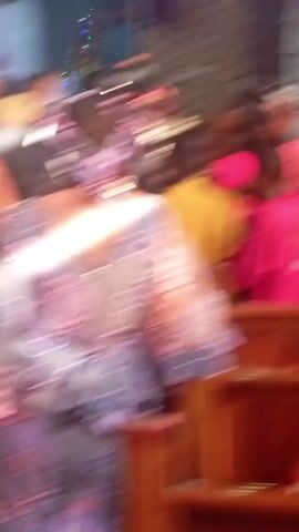 The video is very shaky and blurry. It appears to be taken indoors, possibly in a church or event space. There are people present, including children and adults. The camera moves rapidly, making it difficult to discern specific details about their clothing or activities. Elements of the environment visible include stained glass windows, wooden furniture, and decorative objects. The overall impression is one of a crowded, active space with a potentially celebratory atmosphere.