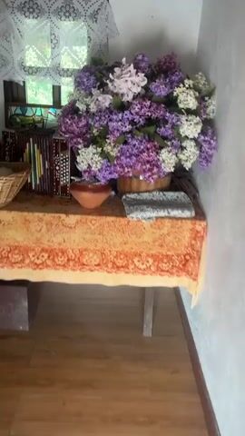 The video pans across a dimly lit room, likely a living space. The first shot focuses on a table covered with an orange, patterned tablecloth. On the table sits a large bouquet of artificial lilacs in various shades of purple and white, housed in a ceramic vase. To the left of the vase, an accordion with decorative inlays is visible, placed next to a stack of books. A small, brown ceramic bowl is also on the table. The camera then pans to the right, revealing a folded grey and white patterned fabric. The perspective shifts to show a window with a white lace curtain. Further panning reveals a heart-shaped decoration on the wall, adorned with white pom-poms, and a framed painting of a landscape. A basket filled with purple onions and some white fiber is also visible, along with a part of a small table or shelf holding other items. The scene transitions to a corner with a black folding chair and a table covered with the same orange tablecloth. A bunch of dried onions hangs on the wall. The camera then moves to another section of the room, where various embroidered shirts and skirts are displayed on hangers and draped on a surface. The overall impression is of a rustic, somewhat cluttered living space possibly adorned with traditional elements.