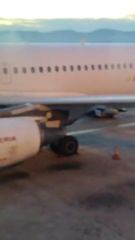 The video shows an Iberia airplane from the window of another airplane on a tarmac. The Iberia airplane has its engine visible, along with the words "IBERIA" on the engine. A yellow vehicle with flashing lights is visible driving on the tarmac. In the background, another airplane is seen landing on the runway with airport lights visible in the distance. A person is speaking in the background.
