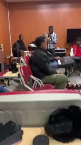 A group of five young men are playing musical instruments in what appears to be a music studio or rehearsal space.  One man is singing into a microphone, using his cell phone. Two men are seated playing keyboards. One man is playing percussion instruments and another is playing bass. The room has wood-paneled walls. A large screen television is mounted on the wall.  A person wearing a black fluffy item is seated near the camera.  Several other chairs are visible, and a guitar is resting on one.