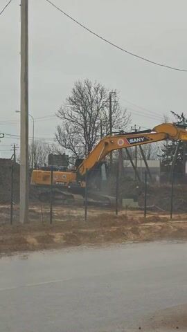 The video shows a yellow Sany excavator operating in a construction site. The excavator is behind a green chain link fence, next to a street. There are piles of dirt on the site. A bare tree and utility poles are visible in the background under an overcast sky.