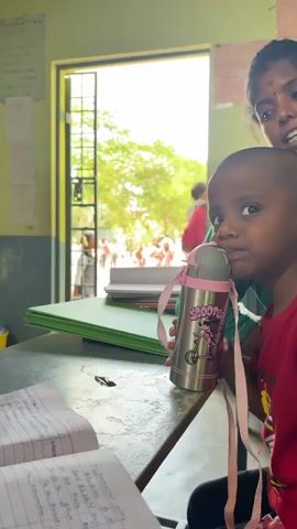 The video opens with a close-up shot of a child with short hair, wearing a red shirt, looking to the right. The child is holding a stainless steel water bottle with a pink strap and a cartoon drawing on it. In the foreground, a notebook with handwritten text is visible on a wooden desk. To the right of the child, a woman wearing a green sari is partially visible, looking downwards. The background shows an open doorway with a metal grate, leading to an outdoor area where several children in red and pink clothing are gathered under trees. The environment appears to be a classroom or a school setting. The video then transitions to a slightly wider shot, focusing on the child and the water bottle, with the background scene remaining the same. The woman on the right is still partially visible. The notebook in the foreground continues to be visible. The video cuts at 00:05.