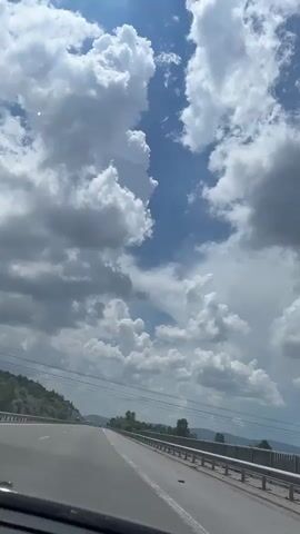 The video shows a view from the driver's seat of a car on a highway. The road ahead is visible, with white lane markers. On the left side of the road, there is a sloping hill covered in trees and vegetation. A guardrail runs along the right side of the road. Overhead, a dramatic sky with large, puffy white clouds against a bright blue sky is visible. Power lines stretch across the sky. The car appears to be in motion.