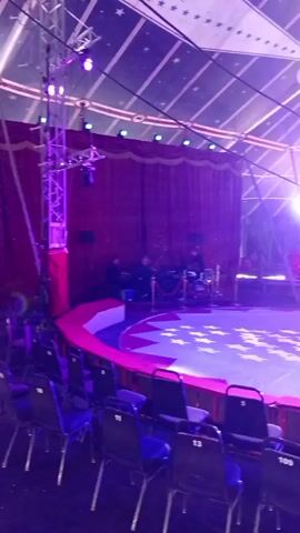 The video shows the interior of a circus tent. In the foreground, there are rows of empty chairs, some with numbers on them. In the middle ground, there is a circular performance area covered with a patterned tarp, and further back, a stage with red curtains and a drum set. The tent ceiling is visible, decorated with a star pattern. Lighting appears to be set up, with spotlights illuminating the performance area. The overall atmosphere suggests the venue is being prepared for a show.