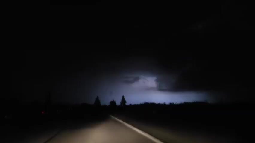 A car drives on a dark road at night, with only the car's headlights illuminating the immediate path ahead. Distant red lights are visible further down the road.