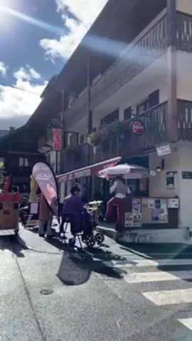 A group of street musicians is playing in front of a building labeled "Bistrot Les Touristes," with a "BAR" sign above. The building has a red awning and a balcony. A drummer in a purple suit sits in a wheelchair. A guitarist stands to the left, and another figure in striped pants stands near the entrance. A promotional banner stands nearby. The street appears to be paved, with a crosswalk in the foreground. The sky is partly cloudy with bright sunlight.